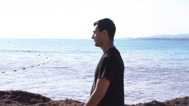 Kumsalda yalnız yürüyen yakışıklı genç adam. Medya. Güneşli bir yaz gününde, deniz kıyısında yavaşça yürüyen siyah saçlı bir erkek..