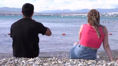 Denizin önündeki taşlı sahilde oturan sevgi dolu çiftin arka görüntüsü. Medya. Balayı kavramı, aşık bir erkek ve kadın güneşli bir yaz gününde kumsalda bir bardak kırmızı şarabın tadını çıkarırlar..