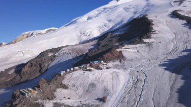 Kar, kış mevsiminde dağların zirvelerini tırmananlar için bir sığınak haline getirdi. Şarjör. Mavi gökyüzü arka planındaki gezginler ve dağcılar için karlı yamaçların ve çeşitli binaların havadan görüntüsü.