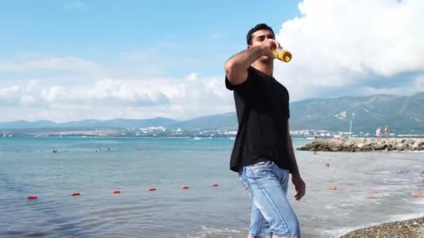 Ung stilig man dricker öl på stranden och molnig himmel bakgrund. Begreppet. Manlig avkoppling på havet stranden framför gröna berg, sommartid och frihet koncept. — Stockvideo