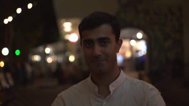 Handsome smiling man winking at someone in front of the camera. Media. Dark haired man in white shirt flirting at the outdoor restaurant terrace.