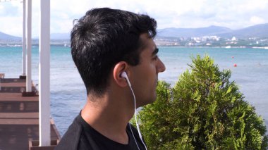 Deniz kenarında müzik dinleyen yakışıklı bir adamın portresi. Medya. Sakin, siyah tişörtlü, kulaklıkla deniz kenarından müzik dinleyen genç adamın yan görüntüsü.