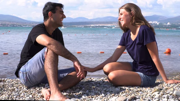 Man och kvinna pratar med varandra framför havet. Media. Två glada smolong vänner sitter tillsammans på en stenig strand under sin semester i en idyllisk natur scen bakgrund. — Stockfoto