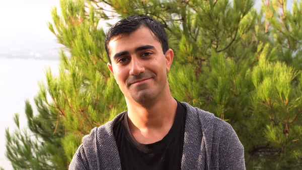 Portret van een knappe lachende man met nat donker haar in een zomerpark. De media. man kijkt in de camera en glimlacht op de achtergrond van groene dennenboom. — Stockfoto