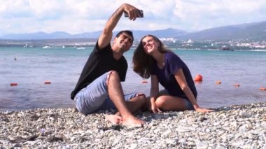 Kumsalda akıllı telefonlarıyla fotoğraf çeken mutlu romantik bir çift. Medya. Kadın ve erkek çakıl taşı kumsalında oturup selfie çekiyorlar..