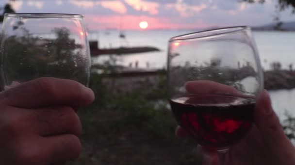 Close up de mãos levantando copos de vinho tinto na praia durante o pôr do sol, celebração ou conceito de data. Mídia. Mãos masculinas e femininas com copos de vinho em um ambiente romântico à beira-mar. — Vídeo de Stock