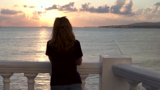 Ung smal tjej i svart t-shirt som står vid havet och lutar sig mot räcket under soluppgången. Media. Bakifrån av en smal kvinna njuter hisnande marint landskap. — Stockvideo