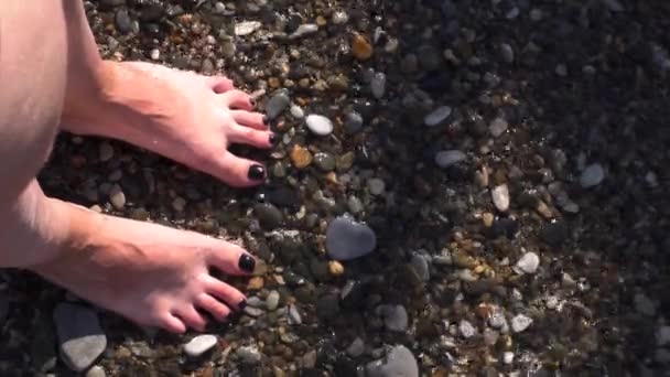 Κοντινή θέα των ποδιών της νεαρής γυναίκας που στέκεται στην ακτή με το κύμα της θάλασσας να φτάνει στα πόδια. Μέσα ενημέρωσης. Καλοκαιρινή δραστηριότητα, όμορφο φόντο διακοπών. — Αρχείο Βίντεο