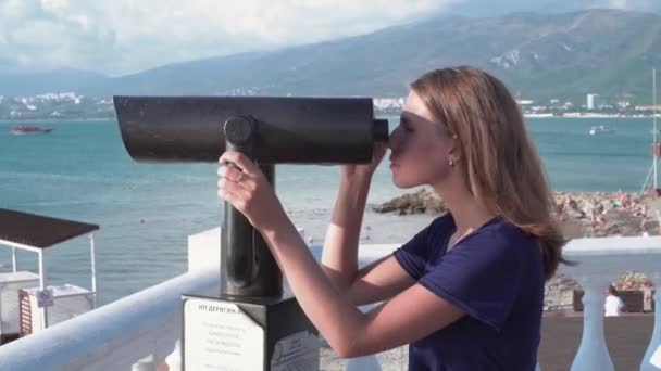 Beautiful woman looking through binoculars on background of sea and mountains. Media. Young woman views seascape with mountains in pair of binoculars on sunny day — Stock Video