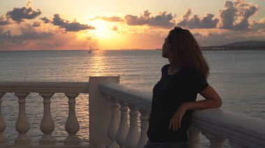 Gün batımında okyanusun derinliklerine bakan sevimli genç bir kadın. Medya. Arkasındaki muhteşem deniz manzarasında kafasına güneş gözlüğü takmış sıska bir kız..