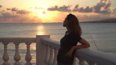 Gün batımında okyanusun derinliklerine bakan sevimli genç bir kadın. Medya. Arkasındaki muhteşem deniz manzarasında kafasına güneş gözlüğü takmış sıska bir kız..