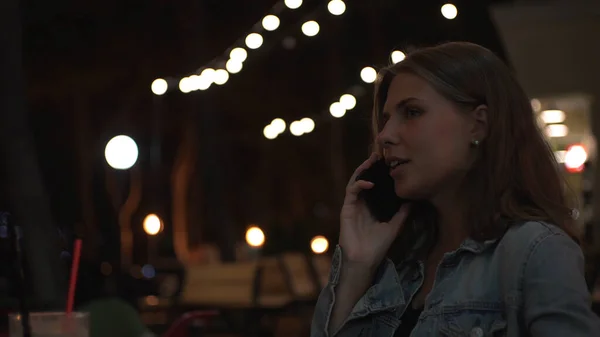 Femeia caucaziană vorbind pe telefonul ei inteligent în timp ce stătea la cafeneaua trotuarului în seara de vară. Media. O tânără femeie cu o ceașcă roșie de cappuccino sau ceai are o conversație telefonică . — Fotografie, imagine de stoc