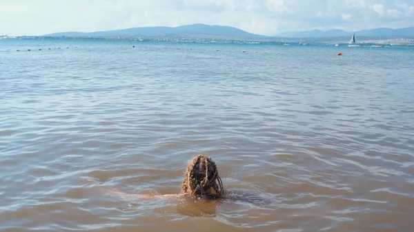 Ung kvinna tar ett dopp i havet. Begreppet. Kvinna på semester simmar i havet med lerigt vatten. Kvinnan simmar i havet på bakgrund av segelfartyg och berg. Semester till sjöss — Stockfoto