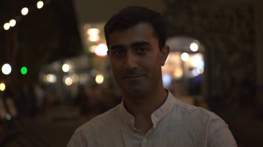 Yakışıklı, gülümseyen bir adam kameranın önünde birine göz kırpıyor. Medya. Beyaz tişörtlü, açık hava restoranının terasında flört eden koyu renk saçlı bir adam..