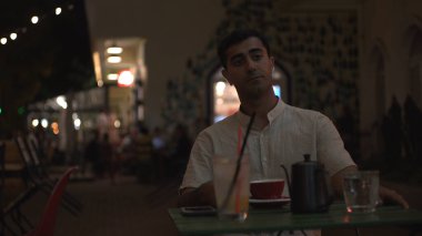 Genç düşünceli adam gecenin geç saatlerinde terasta bir fincan sıcak kahve ya da çayla tek başına oturuyor. Medya. Beyaz tişörtlü, üzgün bir erkek sokak kafesinde yalnız başına birini bekliyor..