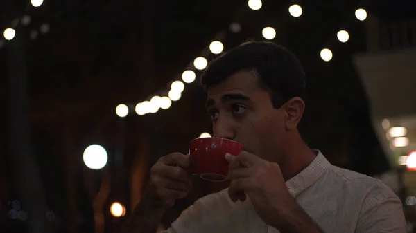 Joven hombre guapo sentado afuera en la cafetería de la ciudad y bebiendo bebida caliente de una gran taza roja. Medios. Hombre bebiendo café en la noche en el cielo negro y fondo de guirnalda. — Foto de Stock