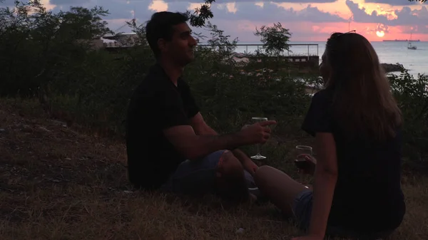Αγαπώντας ευτυχισμένο ζευγάρι πίνοντας κόκκινο κρασί κοντά στην παραλία κατά τη διάρκεια του ηλιοβασιλέματος. Μέσα ενημέρωσης. Άνδρας και γυναίκα συνομιλούν ενώ κάθονται στο έδαφος κοντά στο πράσινο γρασίδι και θάμνους. — Φωτογραφία Αρχείου