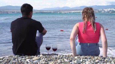 Güneşli bir yaz gününde kıyı kenarında kırmızı şarap kadehleriyle oturan bir çiftin arka görüntüsü. Medya. Siyah gömlekli bir adam ve afro örgülü bir kadın deniz manzarasının tadını çıkarıyor..