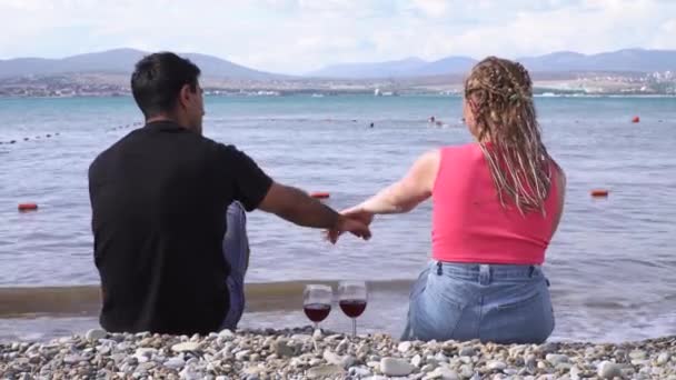 Vista posteriore di uomo e donna che si tengono per mano con due bicchieri di vino rosso in piedi vicino sulla spiaggia di ciottoli. I media. Concetto di luna di miele, donna con trecce e un uomo che si gode il paesaggio marino estivo. — Video Stock