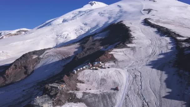 A neve cobriu picos de montanha no inverno com o abrigo para caminhantes. Clipe. Vista aérea da encosta nevada e vários edifícios para viajantes e alpinistas no fundo do céu azul. — Vídeo de Stock