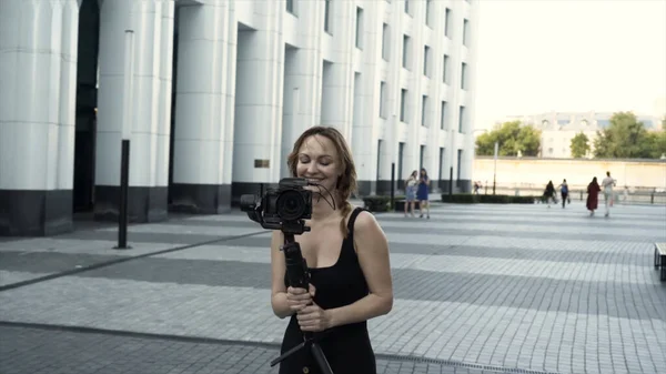 Mulher feliz em férias fotografando com câmera na rua da cidade. Acção. Adulto mulher caucasiana em vestido preto sorrindo e segurando tripé com câmera. — Fotografia de Stock