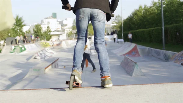 Vista trasera de un joven montando un scooter en un parque de la ciudad. Medios. Hombre cabalgando por el scooter en el campo de deportes en un día soleado de verano. — Foto de Stock