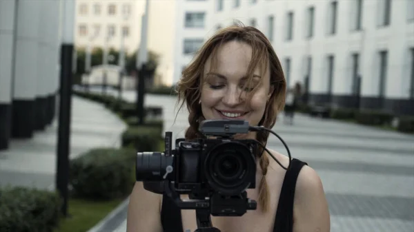 Mulher feliz em férias fotografando com câmera na rua da cidade. Acção. Adulto mulher caucasiana em vestido preto sorrindo e segurando tripé com câmera. — Fotografia de Stock