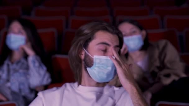 Persone che indossano maschere mediche blu in tempi di epidemia di coronavirus, guardando film al cinema. I media. Ritratto di un uomo stanco che si sfrega gli occhi. — Video Stock