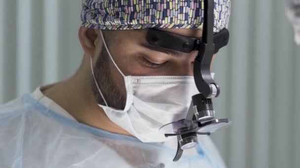 Potret seorang ahli bedah dengan kaca pembesar selama prosedur di ruang operasi. Mulai. Dokter beroperasi dan menggunakan kaca khusus loupes, mengenakan masker bedah dan topi bedah di ruang operasi — Stok Video