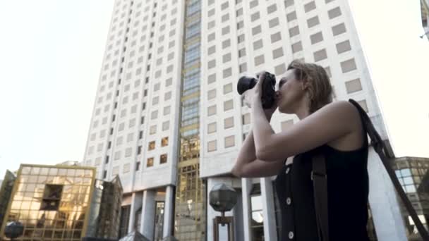 Fotógrafa con cámara profesional. Acción. Mujer joven sosteniendo la cámara en sus manos y tomando fotos en la calle caminar sobre el fondo de un edificio con muchas ventanas y vidrio dorado — Vídeos de Stock