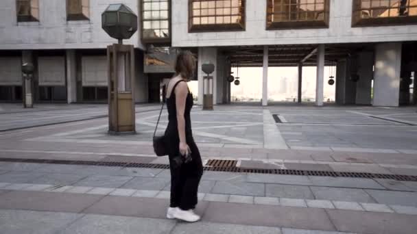 Jeune femme en robe longue noire prend une photo de la ville et sourit. L'action. Belle jeune touriste marchant sur le fond des bâtiments du quartier des affaires avec une caméra professionnelle à — Video