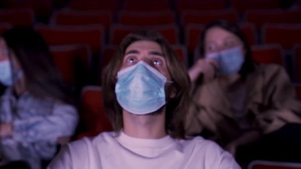 Folk bär blå masker i tider av coronavirus epidemi, titta på film på bio. Media. Porträtt av en ung man som tittar på film med stort intresse. — Stockvideo