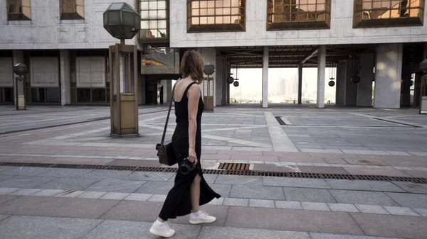 Mulher jovem em vestido preto longo tira uma foto da cidade e sorri. Acção. Bela jovem turista andando sobre o fundo dos edifícios do distrito de negócios com uma câmera profissional em — Fotografia de Stock
