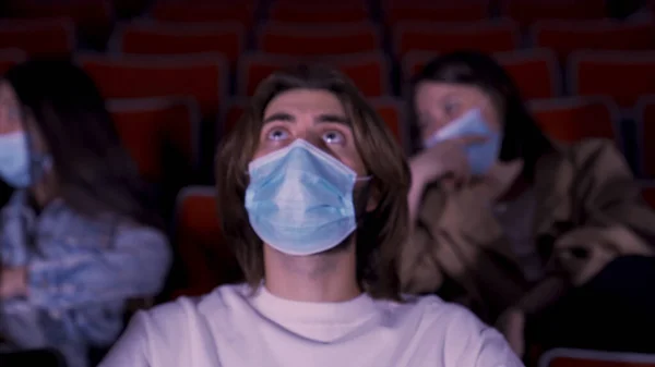 Personas con máscaras médicas azules en tiempos de epidemia de coronavirus, viendo películas en el cine. Medios. Retrato de un joven viendo películas con gran interés. — Foto de Stock