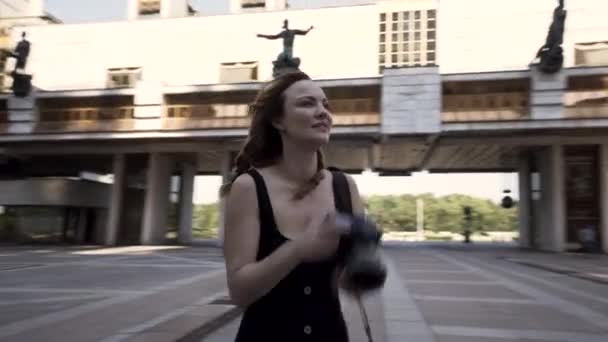 Belle jeune photographe femme prend des photos à l'extérieur d'un quartier d'affaires moderne. L'action. Caméra se déplaçant autour d'une belle femme mince en robe noire avec un appareil photo professionnel dans ses mains. — Video