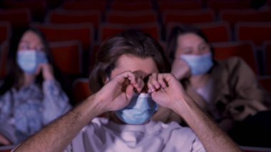 Koronavirüs salgını zamanında mavi tıbbi maskeler takan insanlar sinemada film izliyorlar. Medya. Gözlerini ovuşturan yorgun bir adamın portresi..