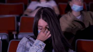 Büyük yeşil gözlü, genç, güzel bir kızın portresi. Sinemada film izliyor, maskesini takıyor ve gözlerini ovuşturuyor. Medya. Sosyal mesafe uygulaması, covid sırasında eğlence kavramı