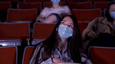 Karantinadaki sinema, koronavirüs salgını güvenlik kuralları, film izlerken sosyal mesafe. Medya. Güzel, uzun saçlı, koruyucu maskeli genç kadınlar oditoryum sıralarında oturuyorlar.