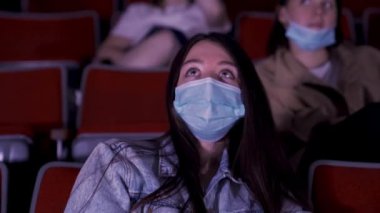 Büyük yeşil gözlü, genç, güzel bir kızın portresi. Sinemada film izliyor, maskesini takıyor ve gözlerini ovuşturuyor. Medya. Sosyal mesafe uygulaması, covid sırasında eğlence kavramı