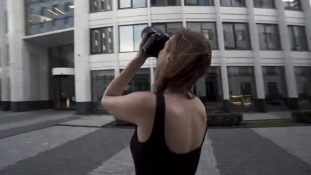 Belle jeune photographe femme prend des photos à l'extérieur d'un quartier d'affaires moderne. L'action. Caméra se déplaçant autour d'une belle femme mince en robe noire avec un appareil photo professionnel dans ses mains. — Video