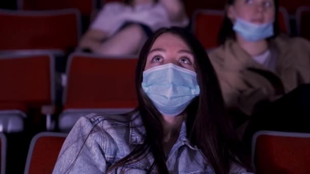 Ritratto di una giovane e stanca bella ragazza con grandi occhi verdi che guarda un film al cinema con maschera medica e si strofina gli occhi. I media. Pratica sociale a distanza, concetto di intrattenimento durante il Covid — Video Stock