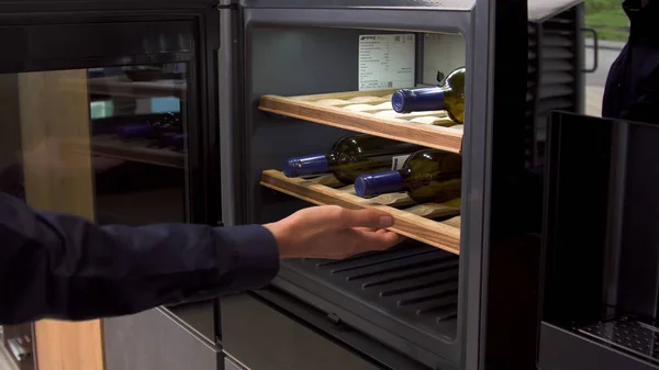 Wijnflessen verkoeling op koelkast houten planken. Huishoudelijke gebruiksvoorwerpen. Sluiten van home use wijnkoeler, de mens opent de deur van een koelmachine. — Stockfoto