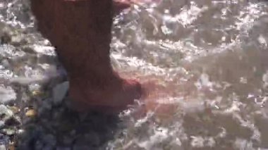 Kayaları ve erkek ayaklarını kaplayan taştan bir sahilde deniz dalgaları. Medya. Deniz kıyısında dikilen adama yaklaş, yaz uykusu ve boş yer kavramı.