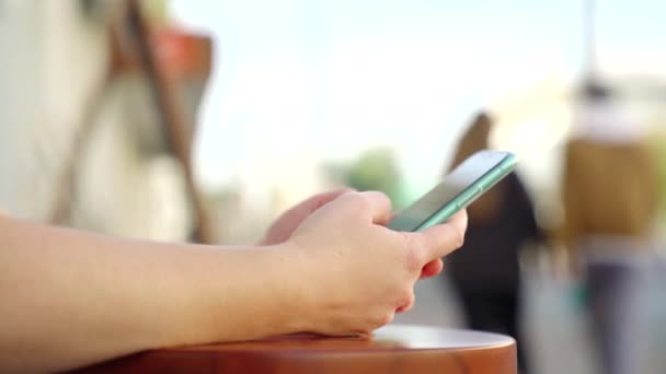 Vista lateral de las manos de mujer sosteniendo un nuevo teléfono inteligente en el fondo borroso de la calle de la ciudad. Medios. Primer plano de una chica enviando mensajes de texto o navegando en su dispositivo electrónico moderno al aire libre. — Vídeo de stock