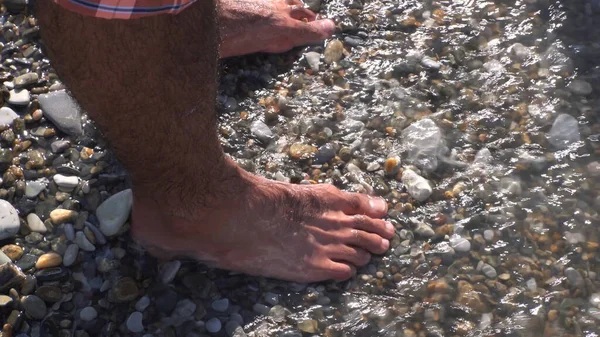 Un mar ondea en una playa de piedra que cubre las rocas y los pies masculinos. Medios. Primer plano del hombre de pie en la orilla del mar, el descanso de verano y el concepto de vacante. — Foto de Stock