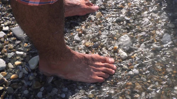 Ein Meer wogt an einem steinernen Strand, der Felsen und Männerfüße bedeckt. Medien. Nahaufnahme eines Mannes, der am Meer steht, Sommerruhe und Leerstandskonzept. — Stockfoto