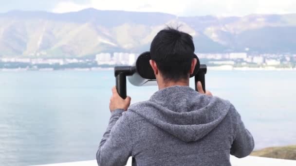 Muž si užívá nádherný výhled. Média. Zadní pohled na mladého cestovatele hledícího dalekohledem s mincemi na pozadí krajiny s mořem a horami.