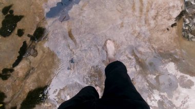 Temiz bir dağ nehrinde, dev taşların üzerinde yalınayak yürüyen bir adam. Şarjör. Seyahat konsepti, erkek bacaklarının ve ayaklarının soğuk akıntıda yürümesinin üst görüntüsü..