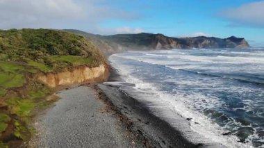 Güçlü okyanus dalgaları, güneşli bir günde yemyeşil kayalıkların yakınındaki siyah bir kumsala çarpar.