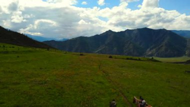 Arka planda dağlar olan yeşil bir çayırda at süren insanların hava manzarası. Medya